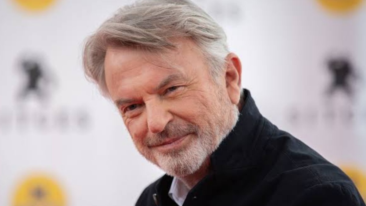 Sam Neill 1 (1) - THEWILL NEWS MEDIA Portrait of a smiling older man with gray hair and a light beard, wearing a dark shirt.
