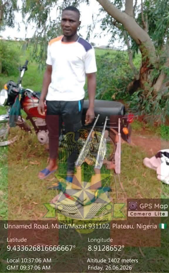 Man standing beside a red motorcycle on a grassy dirt road in a rural Nigerian setting with trees in the background.
