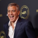 Venice Film Festival to Honor George Clooney with Lifetime Achievement Award Smiling man in a navy suit photographed at an event with a dark backdrop and gold AFI branding behind him.