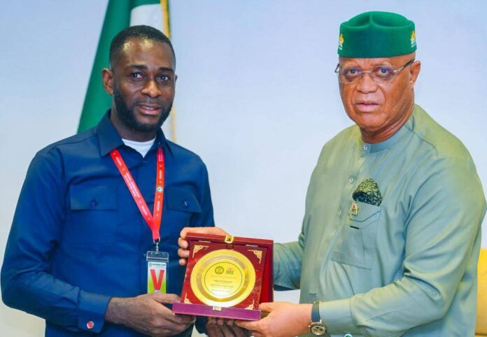 The NUJ Chairman, Akwa Ibom State Council, Comrade Nsibiet John, presenting a plaque to the Governor, Pastor Umo Eno, during the visit to the State Executive Chamber. Two men shake hands and hold a ceremonial award plaque with a gold circle in a red case; one wears a blue shirt with a red lanyard, the other a green traditional outfit and cap.