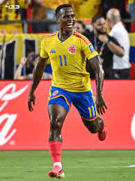 image (1) Colombia national team footballer in yellow jersey with number 11 sprinting on the pitch during a match, fans in the stands behind him.