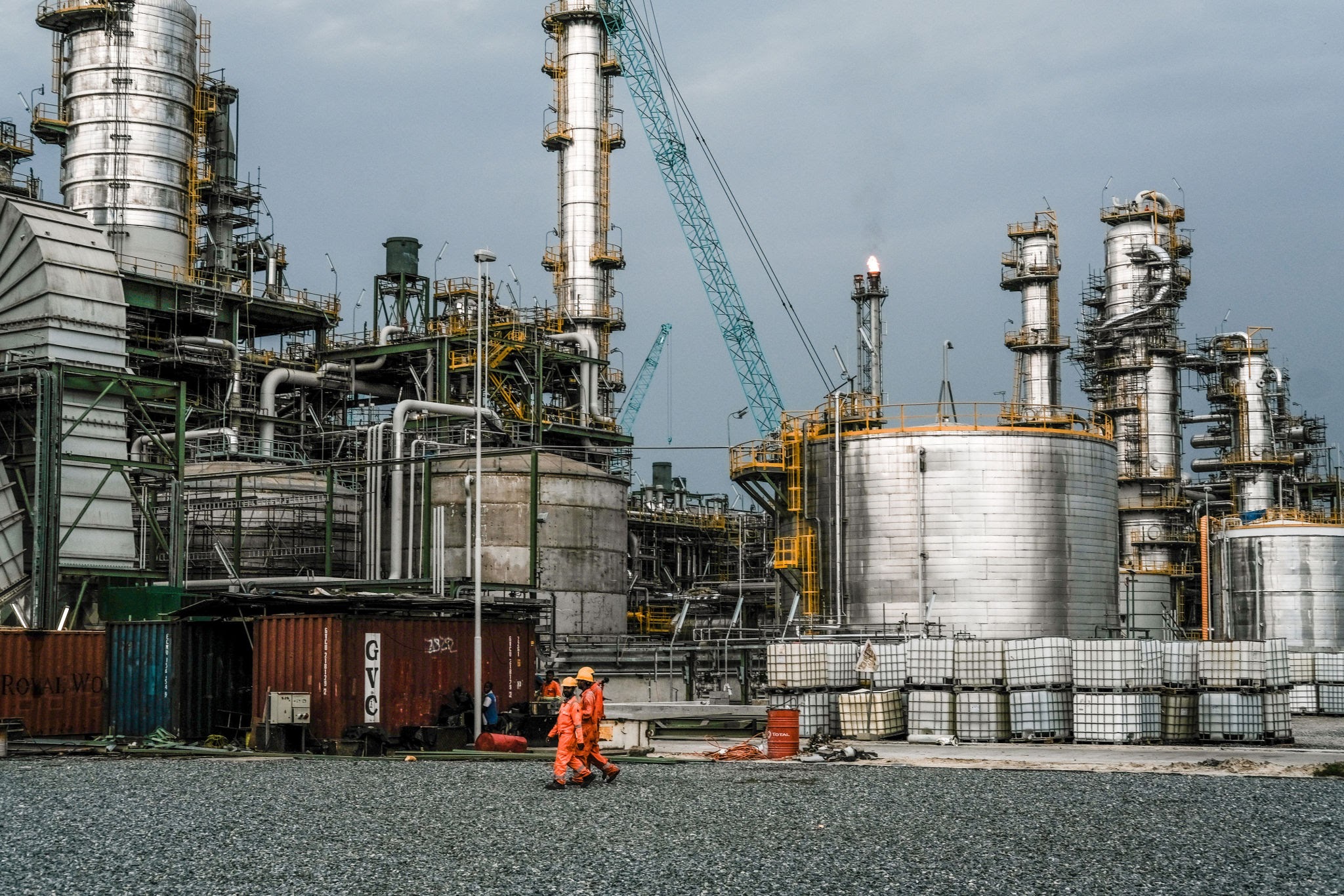 Shipping docks and tankers handling crude oil near Lagos. Source: Dangote Refinery Port - THEWILL NEWS MEDIA Industrial refinery with tall metal towers, pipes, and cranes; three workers in orange safety suits walk in the foreground.