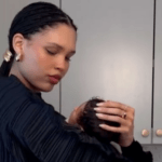 Model Nara Smith Reveals 2-Year-Old Daughter’s Cancer Diagnosis Woman cradling a baby’s head while styling the child’s hair in a kitchen setting, with cabinets in the background.