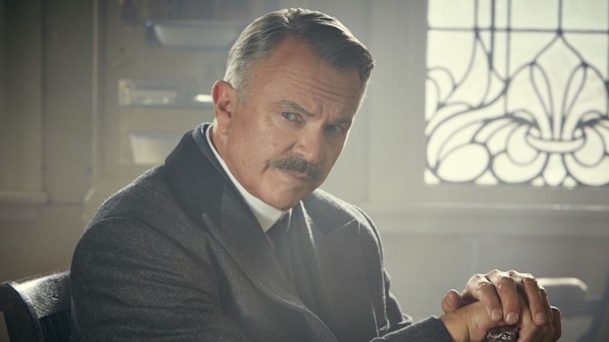 sam neill 3 - THEWILL NEWS MEDIA Older man in a suit sits at a desk with hands clasped, looking intently at the camera in a vintage, sunlit room