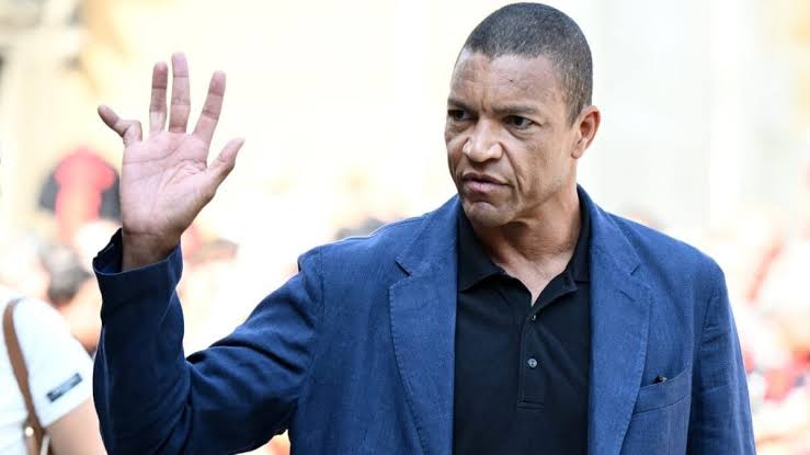 DIDA - THEWILL NEWS MEDIA Man in a blue blazer and black polo raises his right hand in a wave, outdoors, with a focused expression.