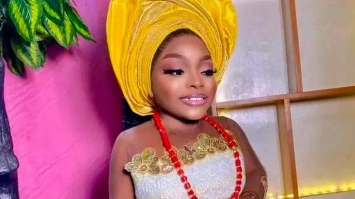 Ajara Lasisi - THEWILL NEWS MEDIA Woman wearing a bright yellow headwrap (gele) with a white lace dress and red bead necklace, posing against a pink wall.