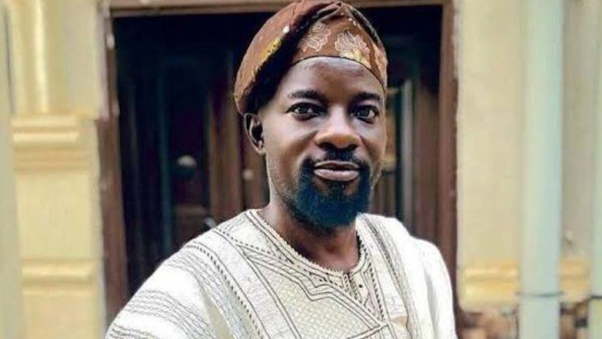 Bamidele Okemesi - THEWILL NEWS MEDIA Portrait of a man in traditional Nigerian agbada and brown headwrap standing outdoors beside a doorway