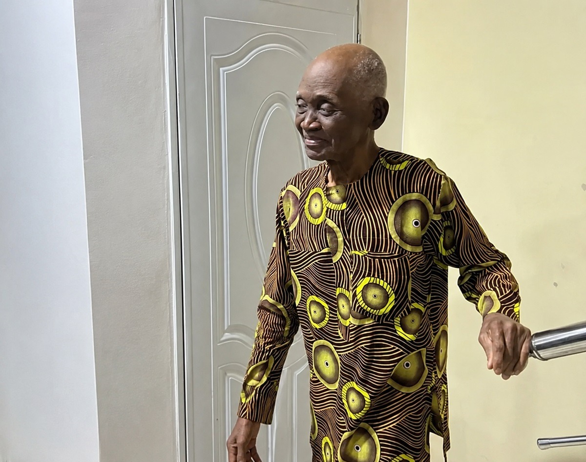 Davis Offor Clarus - THEWILL NEWS MEDIA Elderly man with a bald head and a brown-yellow patterned shirt uses a handrail beside a pale door, smiling slightly on a sunny day.