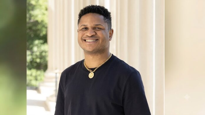 Jerry Falade Smiling man in a black shirt with a gold chain stands beside columned architecture outdoors.