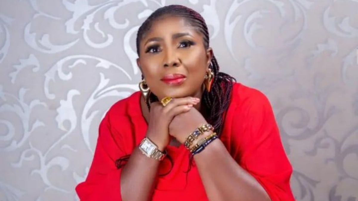 Oby - THEWILL NEWS MEDIA Woman in a red blouse rests her chin on clasped hands, wearing gold jewelry, with a decorative gray backdrop.