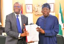 Tinubu Receives ICPC Report On Fake Presidential Agency Two men in an office exchange a document; one in a gray suit and blue tie, the other in blue traditional attire, smiling.",
