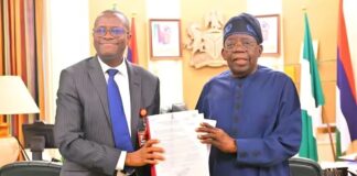 Tinubu Receives ICPC Report On Fake Presidential Agency Two men in an office exchange a document; one in a gray suit and blue tie, the other in blue traditional attire, smiling.",