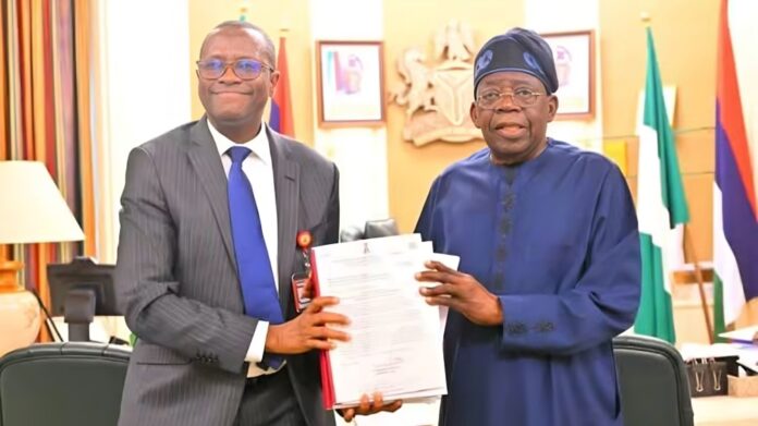 Tinubu Receives ICPC Report On Fake Presidential Agency Two men in an office exchange a document; one in a gray suit and blue tie, the other in blue traditional attire, smiling.",
