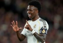 Vinicius Jr Ends Arsenal Transfer Talk By Signing Long-Term Real Madrid Deal Soccer player in Real Madrid jersey with captain's armband celebrating, smiling and gesturing with hands.